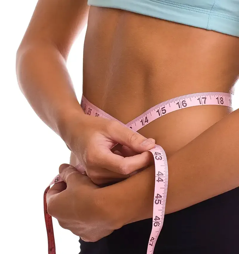 A woman in a crop top measuring her waist with a pink tape measure.